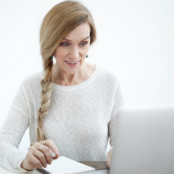 Emotional excited mature woman with long braid waiting at desk with empty copybook, smart phone and tea in paper cup, reading good news online, looking at laptop screen, raising her eyebrows