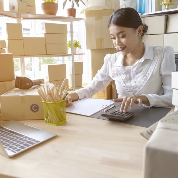Asian woman entrepreneur using calculator with pencil in her hand, calculating financial expense at home office,online market packing box delivery,Startup successful small business owner,SME, concept