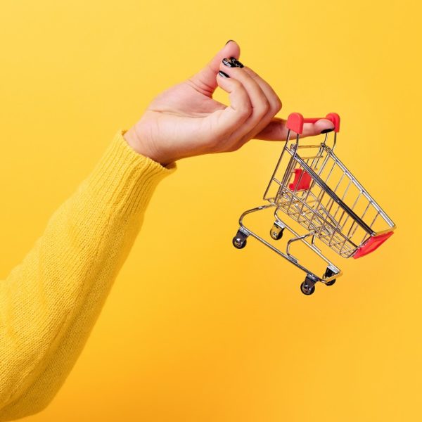 Buying things at market shops concept. Woman hand holding small tiny shopping cart trolley over trend yellow background
