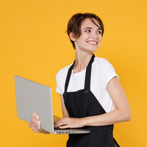 Smiling young female woman 20s barista bartender barman employee in white t-shirt apron posing working on laptop pc computer looking aside isolated on yellow color wall background studio portrait.