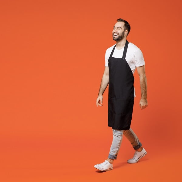 Full length young confident fun man 20s barista bartender barman employee in black apron white t-shirt work in coffee shop walk go isolated on orange background studio. Small business startup concept.