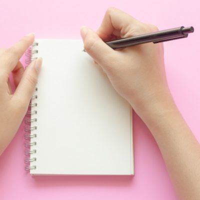 woman's hands writing in empty notebook at the pink desk. Flat lay top view.