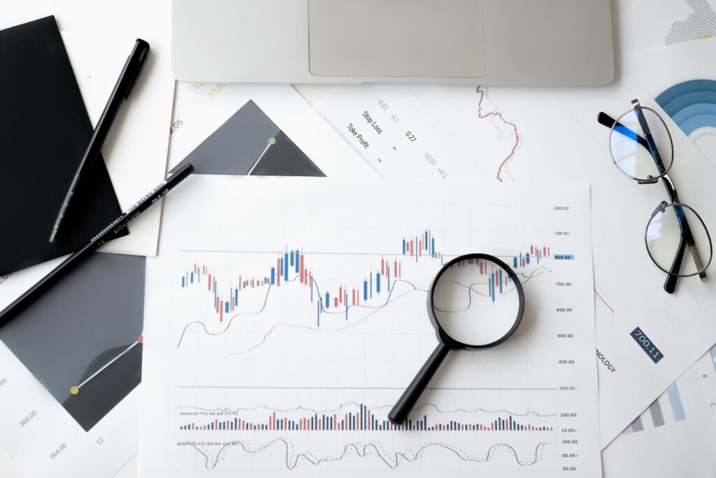 A top-down view of a desk featuring a laptop, glasses, pens, and printed financial charts with a magnifying glass held over a specific data point. Image from